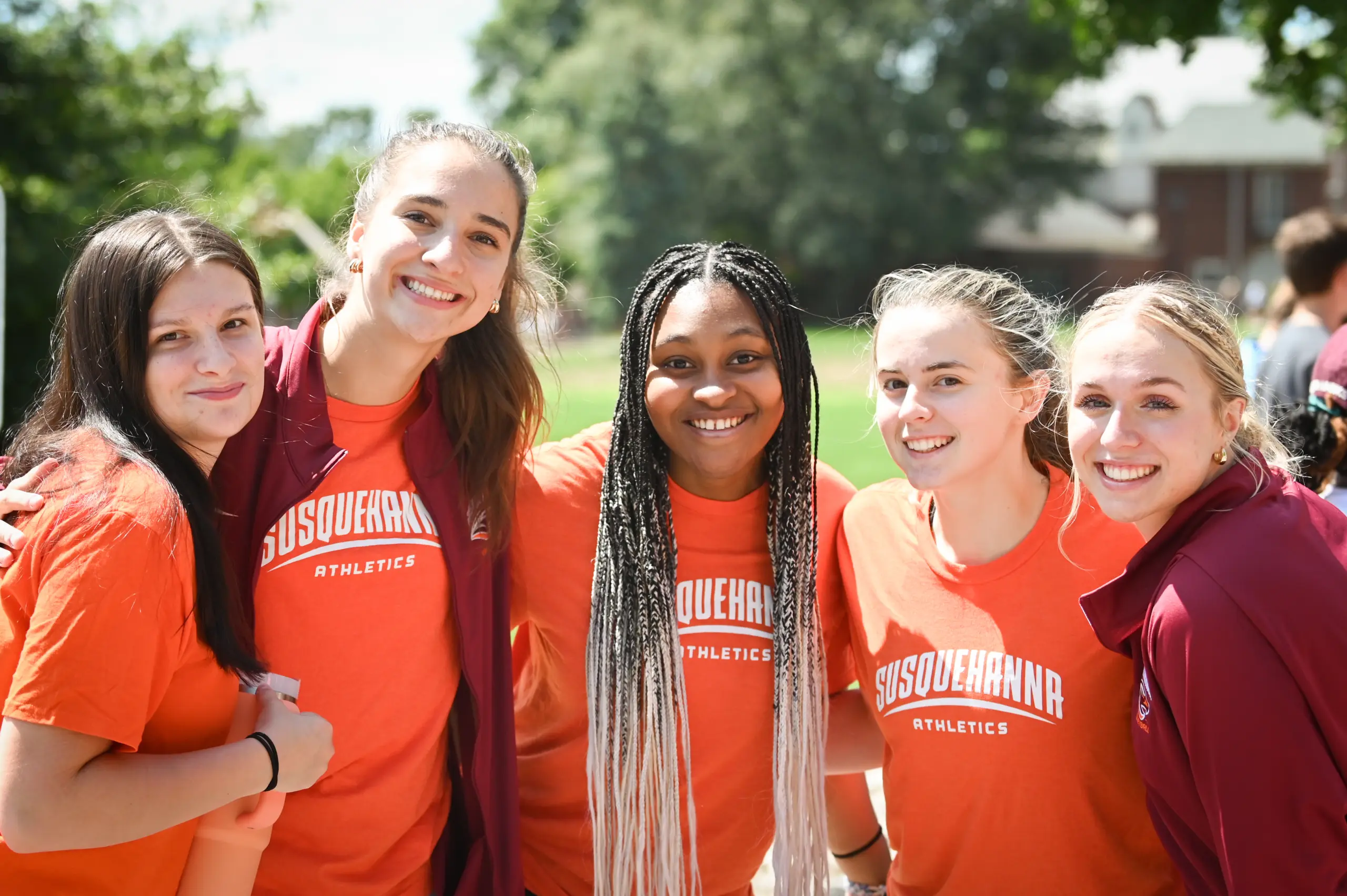 Five smiling young women wearing orange "Susquehanna Athletics" shirts stand close together outdoors on a sunny day, representing Susquehanna Policies, with trees and grass in the background.