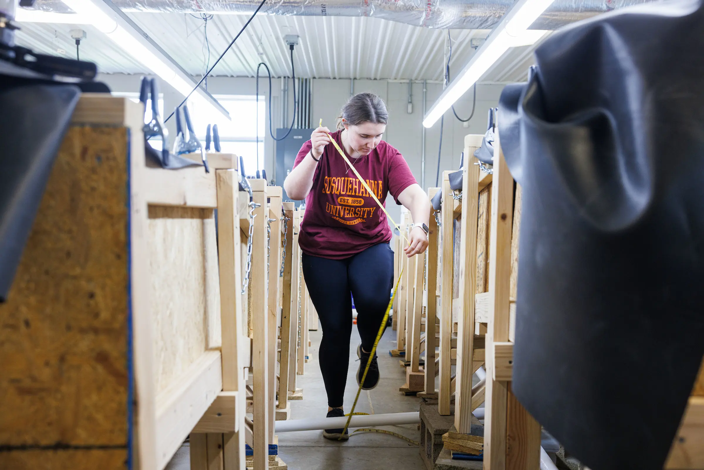 A person in a red university shirt stands in a workshop, holding yellow measuring tape among rows of wooden frames under bright lights.