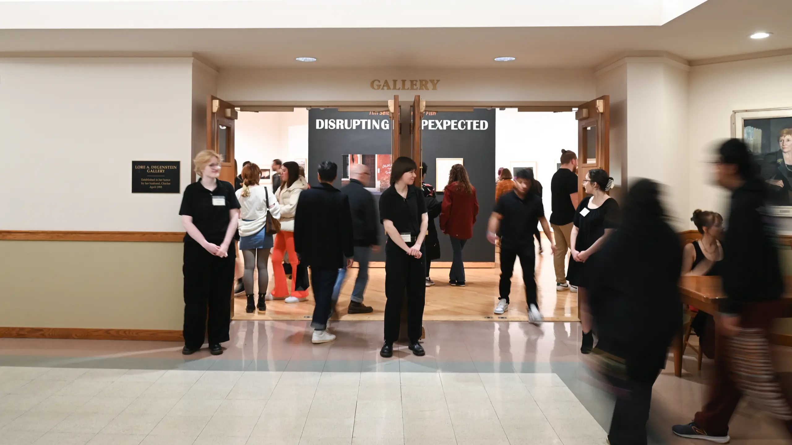 A group of people stand and walk near the entrance of an art gallery titled "Disrupting the Expected." Several staff members in black attire are by the doorway, as visitors—reflecting vibrant campus life—move around inside and outside the gallery.