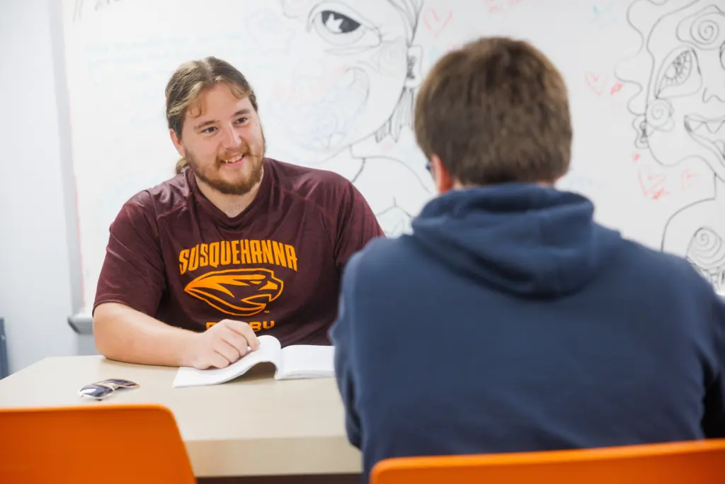 Two people sit across from each other at a table, one wearing a "Susquehanna" shirt and smiling while talking. An open book and phone are on the table, and colorful doodles are on the whiteboard behind them.