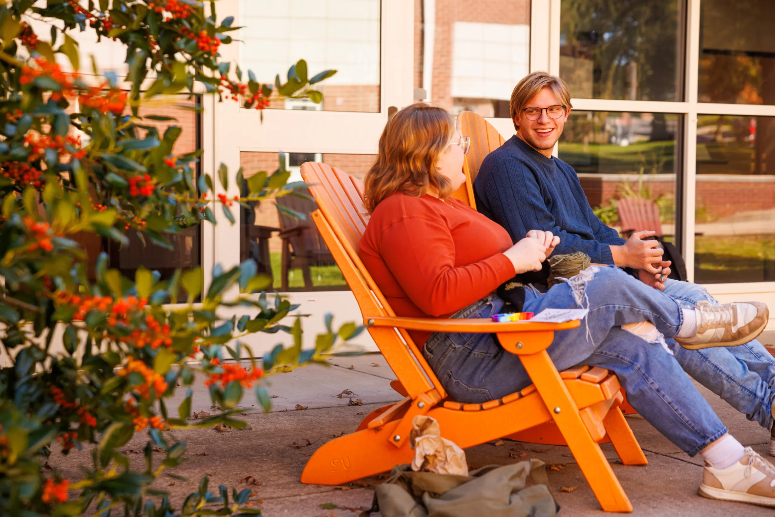 Two people sit on orange outdoor chairs, smiling and talking near a window. One wears a red sweater and glasses; the other has a blue sweater. Green bushes with red berries are in the foreground, adding charm to this scene about friendship.