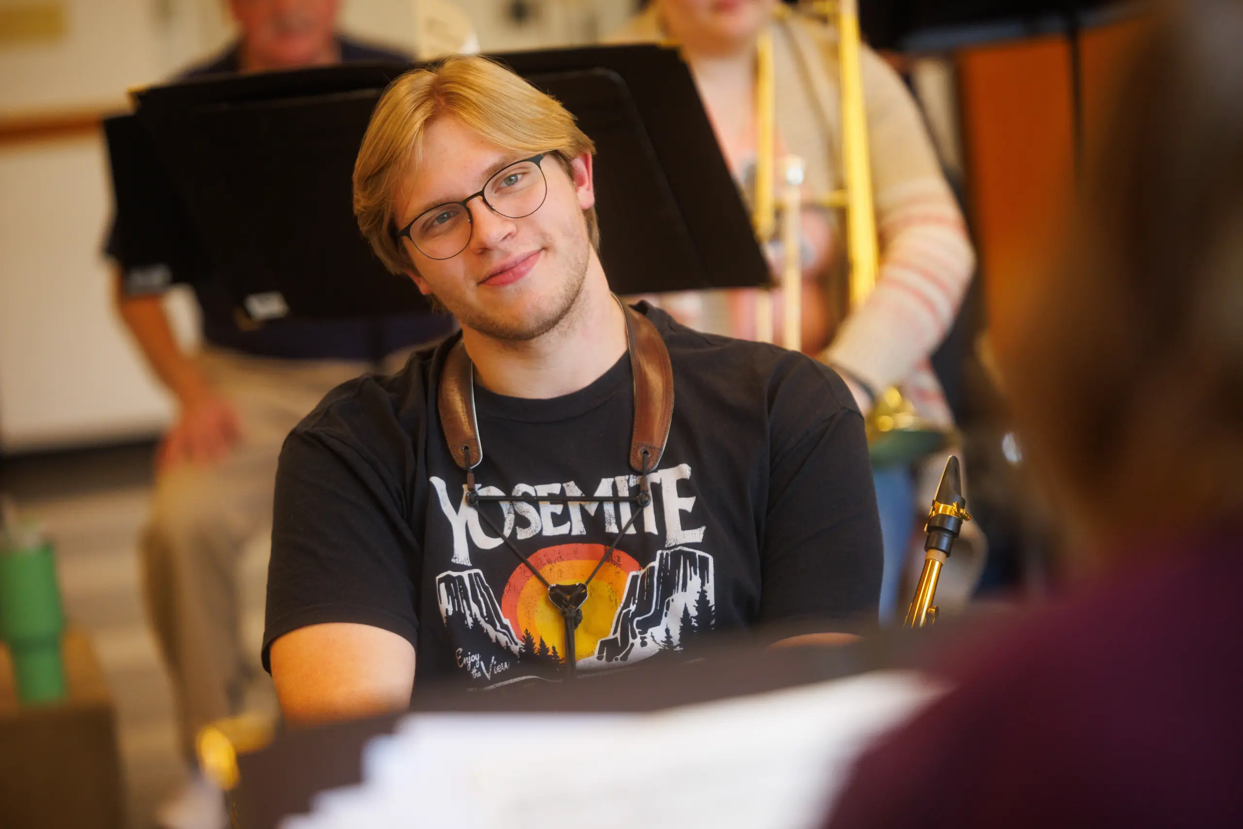 A young man with blond hair and glasses, wearing a "Yosemite" T-shirt, saxophone strap, and an inviting smile, sits in a music rehearsal room where musicians apply their talents in the background.