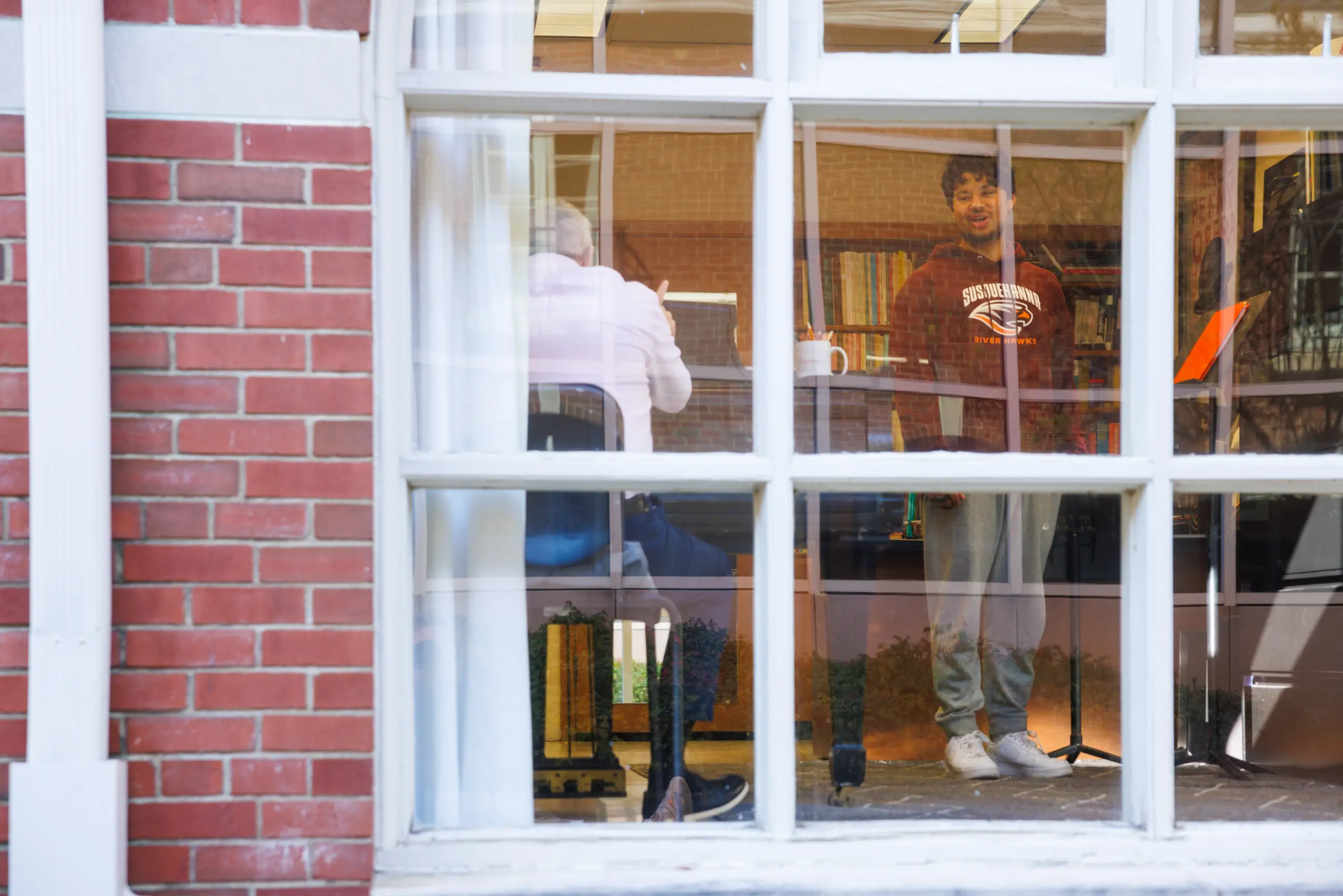 A young man stands and sings near a music stand inside a building, seen through a large window. Another person sits facing him, partially obscured. Bookshelves and warm lighting are visible in the background, reflecting Susquehanna Policies’ community values.