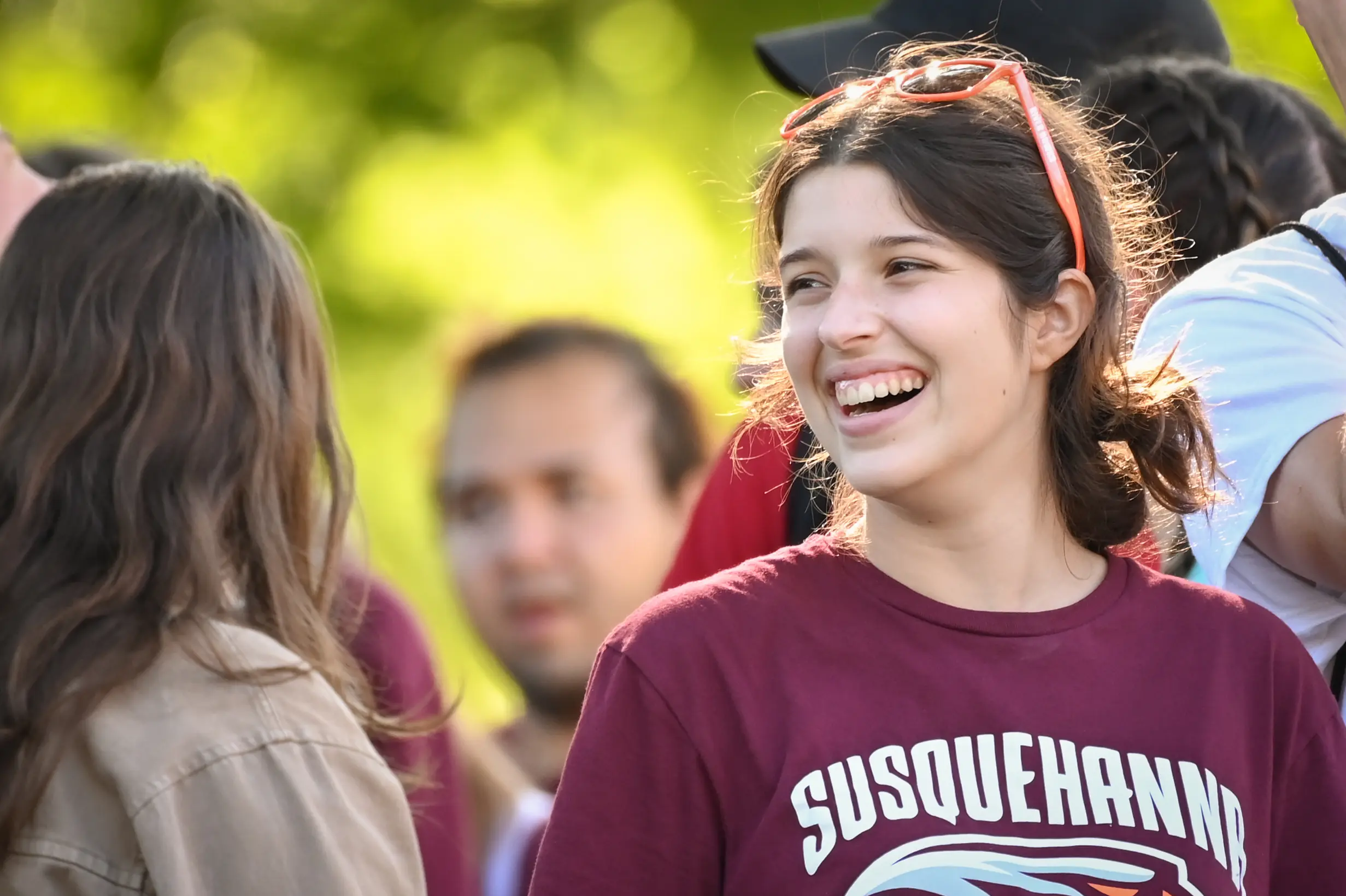 A young woman sporting a maroon "Susquehanna" shirt and sunglasses on her head smiles while chatting about academics with another person outdoors, with blurred people and greenery in the background.