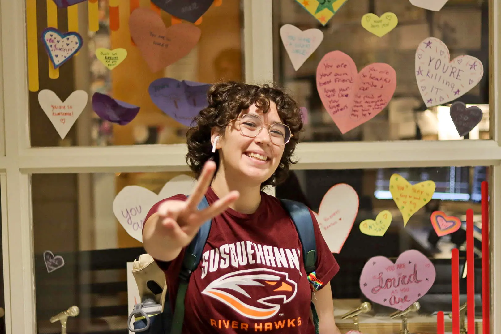 A smiling person wearing glasses and a Susquehanna University River Hawks shirt flashes a peace sign in front of a window decorated with colorful, handwritten heart-shaped notes.