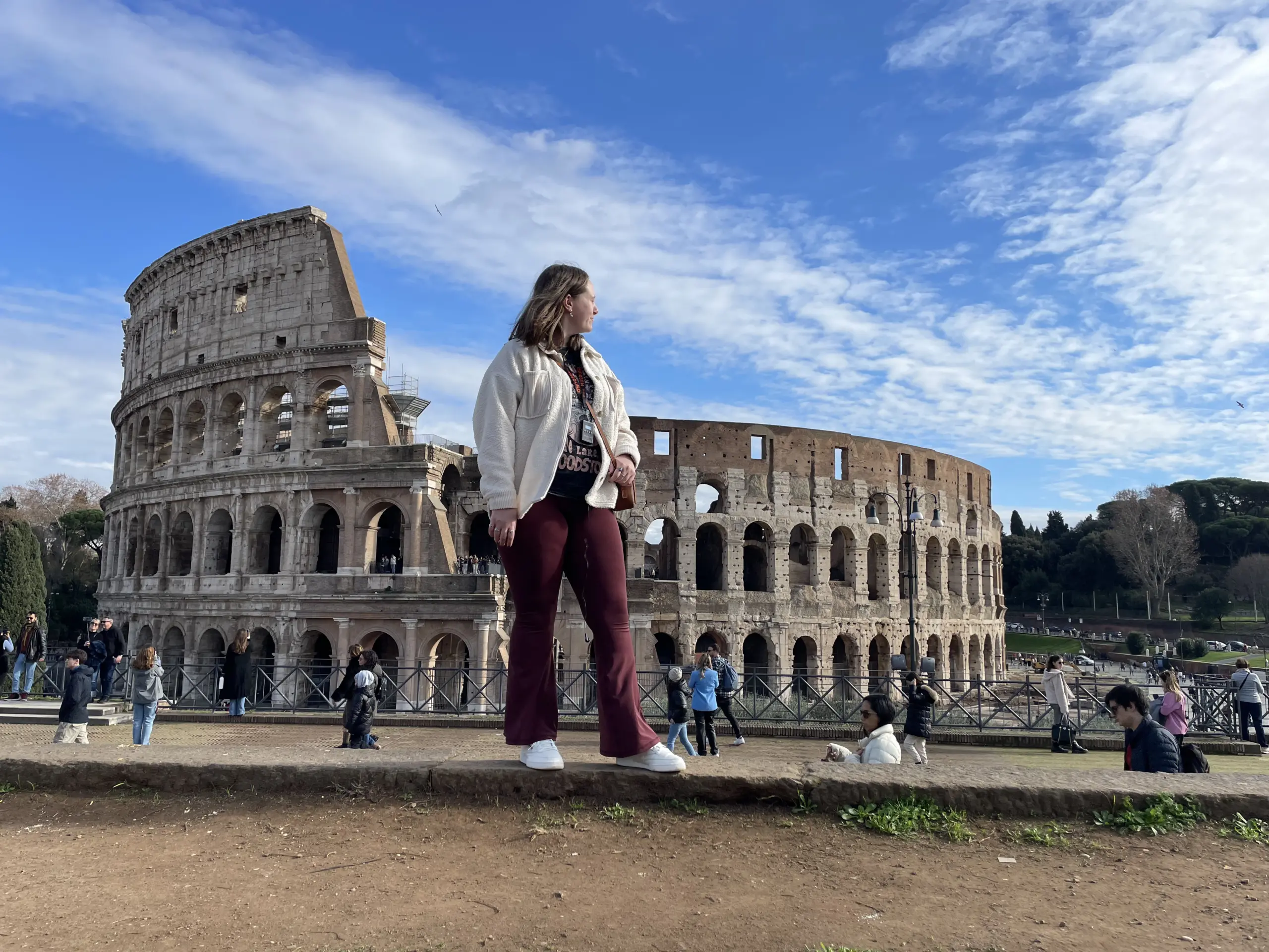 A woman stands on a ledge in front of the Colosseum in Rome, wearing a light jacket and maroon pants. Academics and tourists alike walk and gather around the historic structure under a partly cloudy sky.