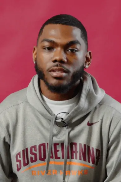 A young man with a trimmed beard wears a gray "Susquehanna River Hawks" hoodie, embodying campus life as he sits against a solid pink background, looking toward the camera.