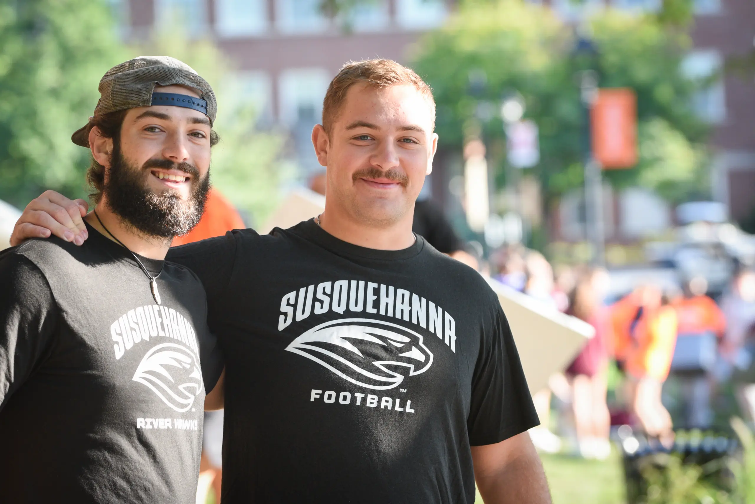 Two young men wearing "Susquehanna Football" t-shirts smile outdoors, one arm around the other’s shoulder. The lively, blurred background hints at a campus event celebrating community and Susquehanna policies.