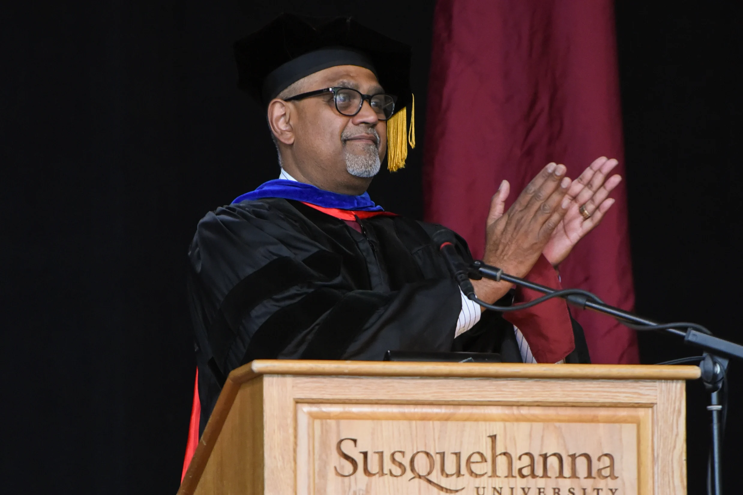 A person in academic regalia stands at a podium with "Susquehanna University" on it, clapping. A flag and a fern are visible in the background.