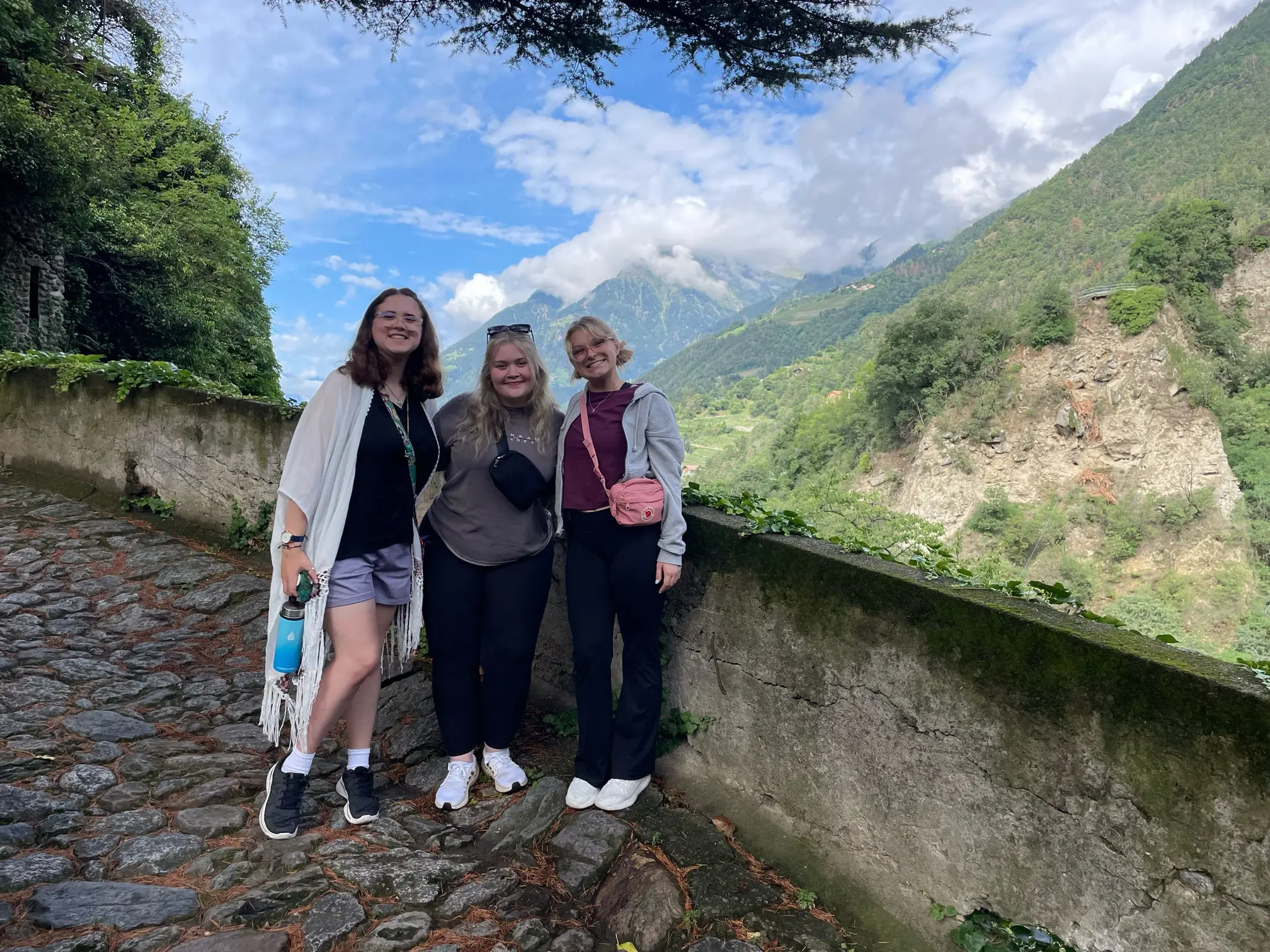 Three women, possibly academics, stand smiling on a cobblestone path bordered by a stone wall, with lush mountains and clouds in the background on a bright, partly cloudy day.