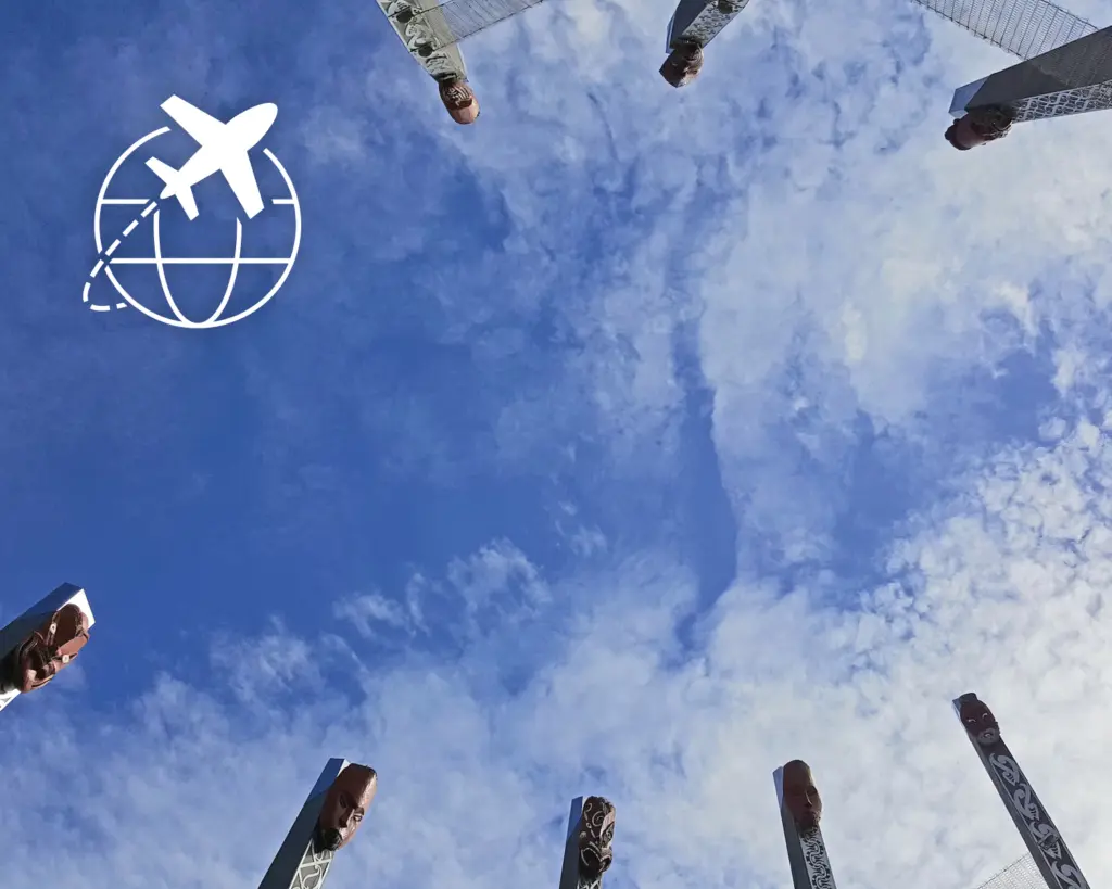 Photo of tall carved pillars with faces, arranged in a circle and viewed from below against a blue sky with clouds; an "About" airplane and globe icon is overlaid on the upper left side.