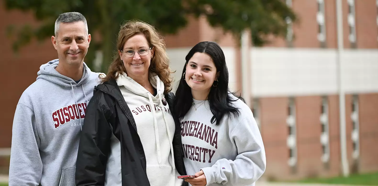Three people stand outside, smiling at the camera. All are wearing Susquehanna University sweatshirts. There are trees and a brick building in the background.