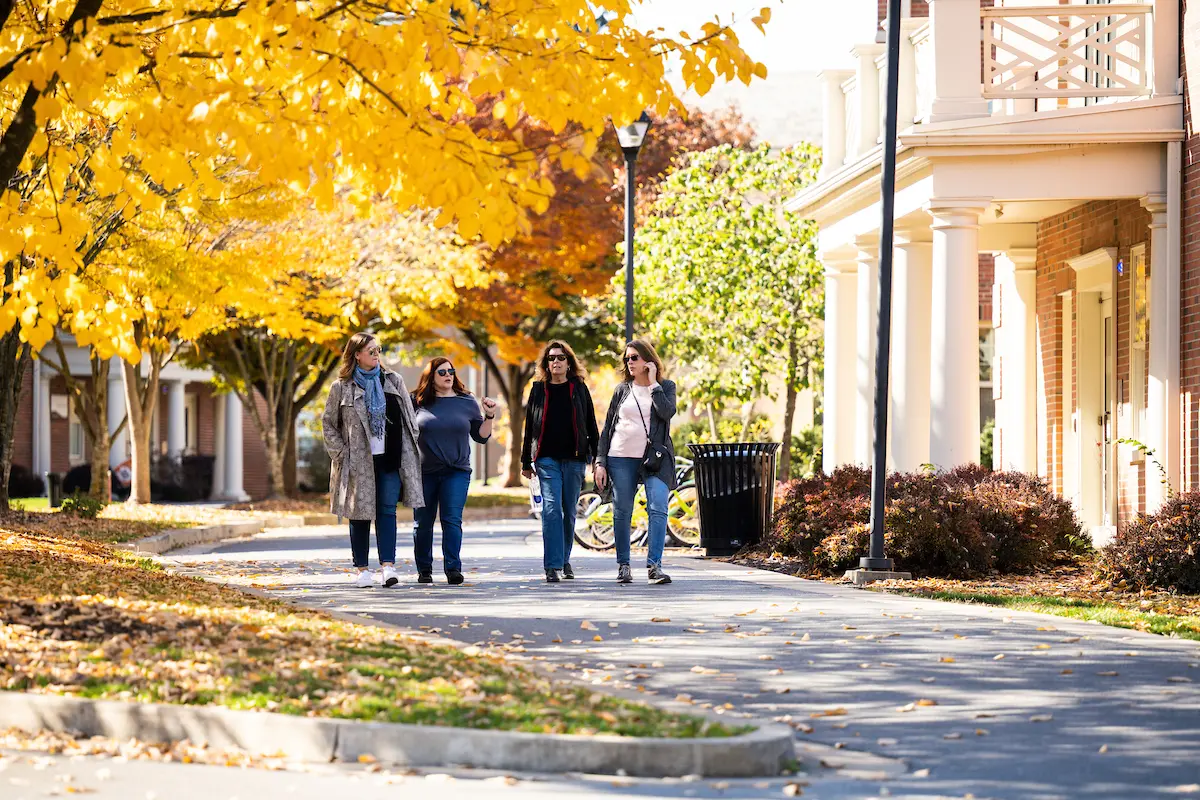 Four women walk together on a tree-lined path with autumn leaves, next to buildings with columns, enjoying a sunny day.