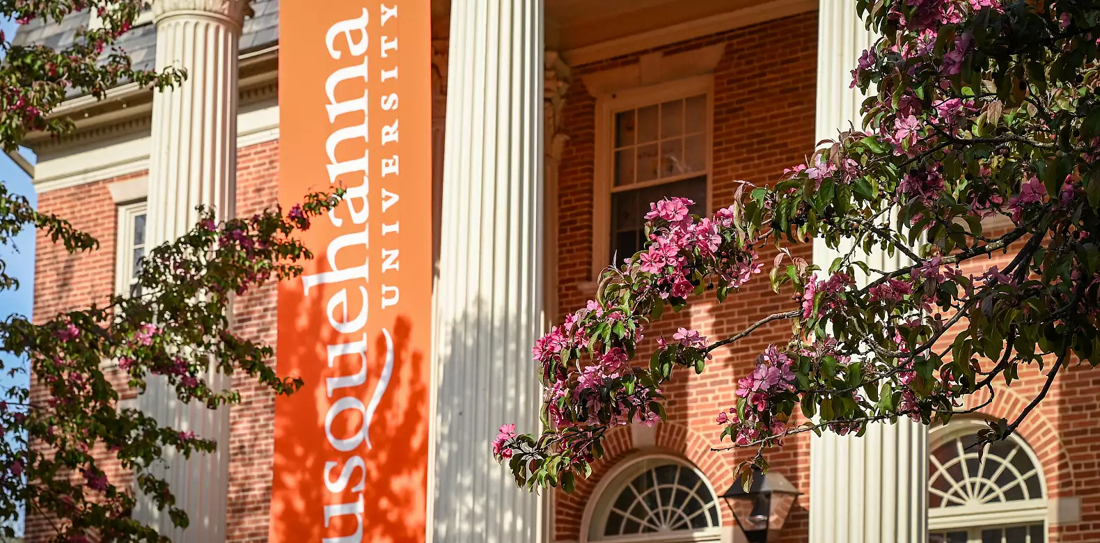 A large orange banner reading "Susquehanna University" hangs between columns of a brick building, with pink blossoming tree branches in the foreground.