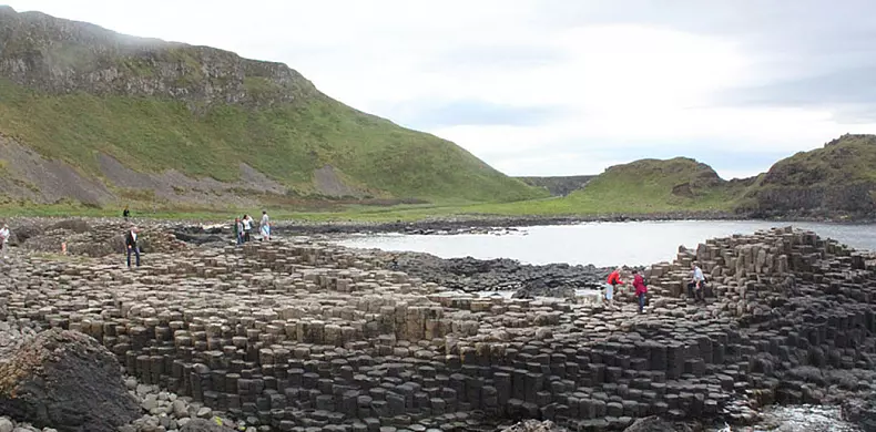 People walk across the Giant’s Causeway in Northern Ireland, a natural formation of interlocking, hexagonal basalt columns by the sea, with green hills in the background under a cloudy sky.