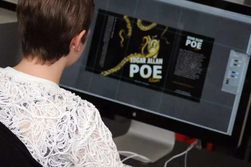 A person with short hair and a white lace top works on a computer, designing a book cover for "Edgar Allan Poe" that features gold and black colors and a graphic of a snake.