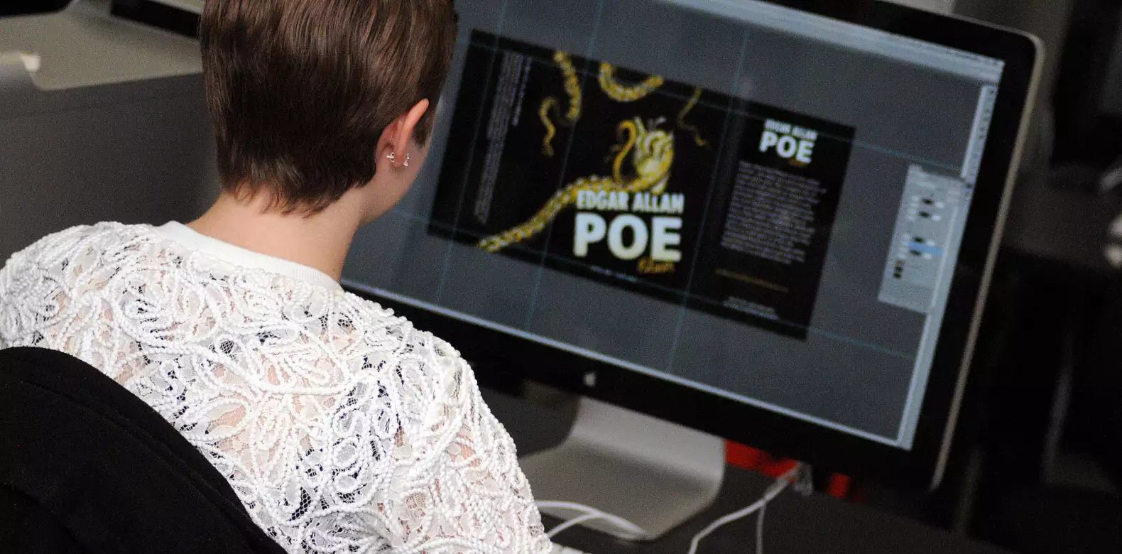 A person with short hair and a white lace top works on a computer, designing a book cover for "Edgar Allan Poe" that features gold and black colors and a graphic of a snake.