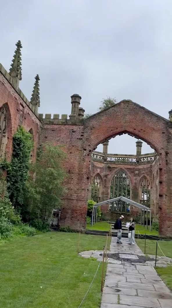 Two people walk along a stone path inside the ruins of a large, roofless brick building with arched windows and ivy-covered walls, evoking an atmosphere where academics might have once gathered, surrounded by green grass under a cloudy sky.