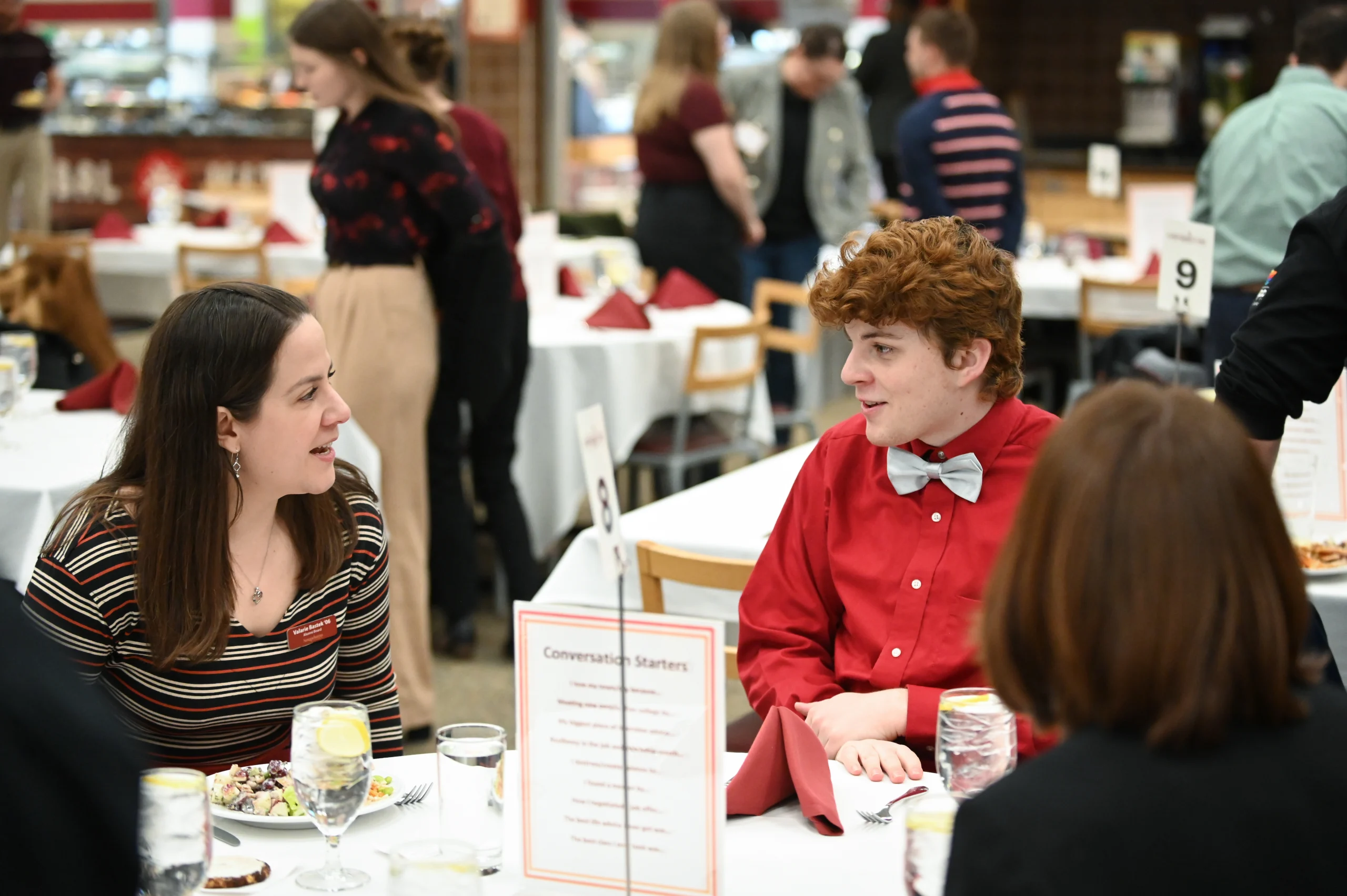 Two people sitting at a table, having a conversation in a dining area. They are surrounded by other guests engaged in discussions.