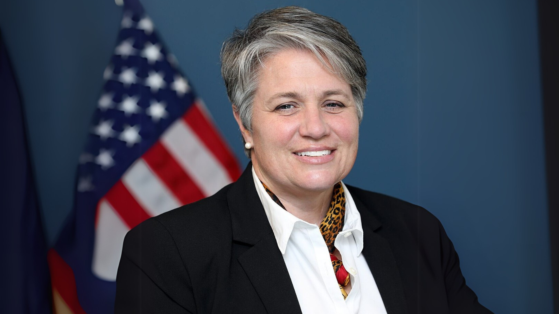 Person in a suit with short gray hair smiles, standing in front of an American flag.