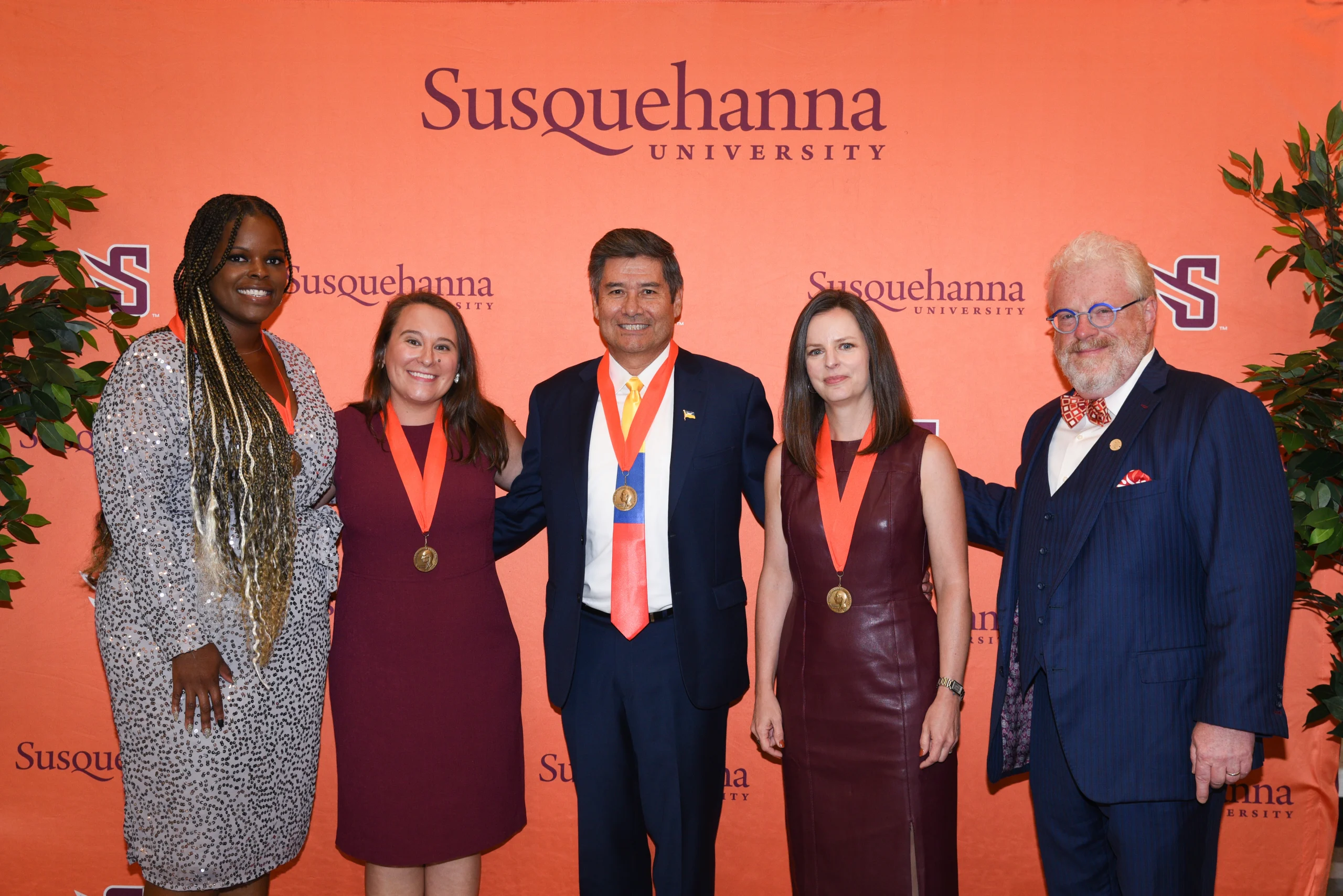 Five people wearing medals stand in front of an orange backdrop with "Susquehanna University" text. Four are smiling, with one man in a blue suit and others in formal attire.